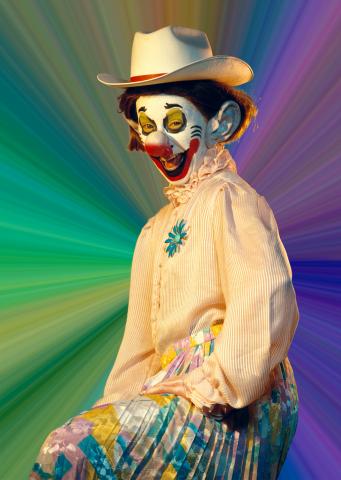 Ein lachender Clown in Bluse, Rock und Cowboyhut sitzt vor einem bunten Hintergrund