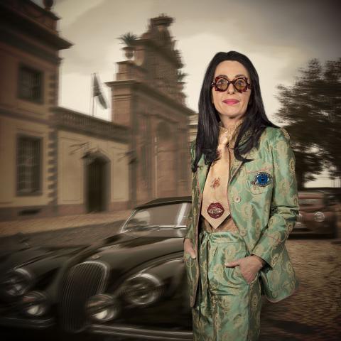 Eine Frau (Cindy Sherman) steht vor einem alten Auto. Sie ist in einem seidig glänzenden Anzug gekleidet und trägt eine auffällige Brille