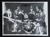 Foto mit zehn Männern in Anzügen. Diese haben sich teilweise in einem Stoffband (Klopapier?) eingewickelt.