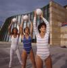 EIn Foto mit drei Frauen, die in Bodysuits aus den 90ern jeweils einen Gymnastikbal über ihren Kopf halten, sie stehen vor dem grünen Eingang der Neuen Staatsgalerie und blauem Himmel.